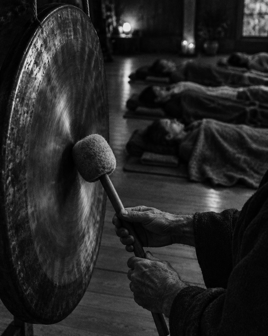 Handen slaan met een vilten mallet een grote bronzen gong aan, op de achtergrond liggen mensen op matjes